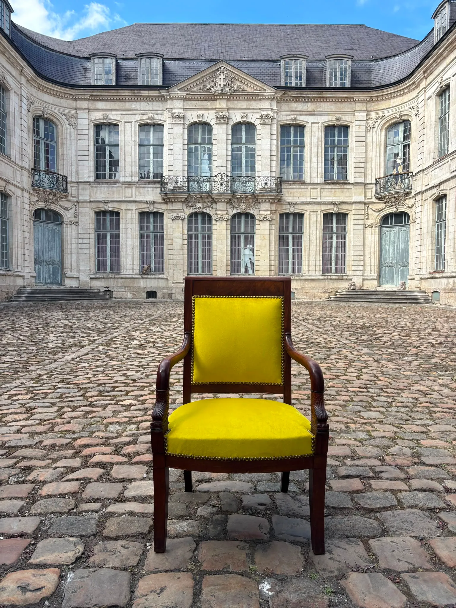 Fauteuil en acajou recouvert d’un velours jaune et clouté laiton, posé sur les pavés d’une cour classique