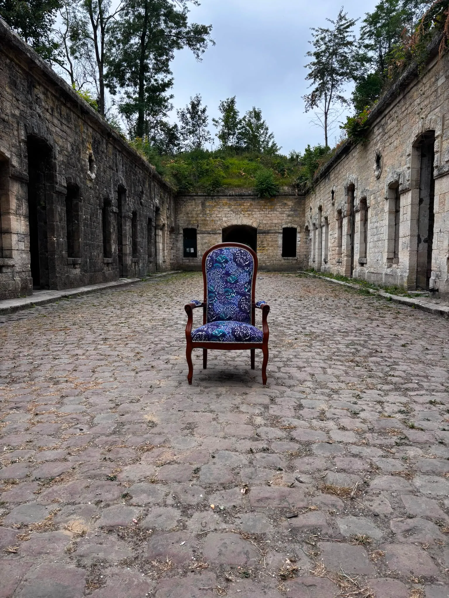 Le fauteuil Voltaire bleu photographié de face au centre d’une cour pavée en pierre