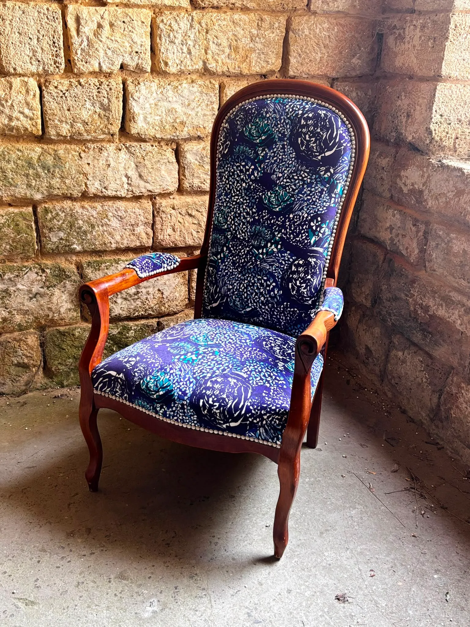 Fauteuil Voltaire habillé d’un imprimé floral bleu et turquoise, clous décoratifs, adossé à un mur de pierre